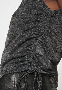 Top in maglia nera e metallica con arricciature laterali e dettagli con cordoncino; presenta una finitura testurizzata e una silhouette aderente.