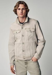 Beige spijkerjack met een knoopsluiting aan de voorkant, twee borstzakken en een rechte snit, gecombineerd met bijpassende lichtgekleurde jeans.
