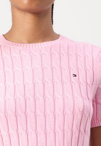 Pull en tricot rose clair à manches courtes, présentant un motif de tricot en relief. Un petit logo est visible sur le côté gauche.