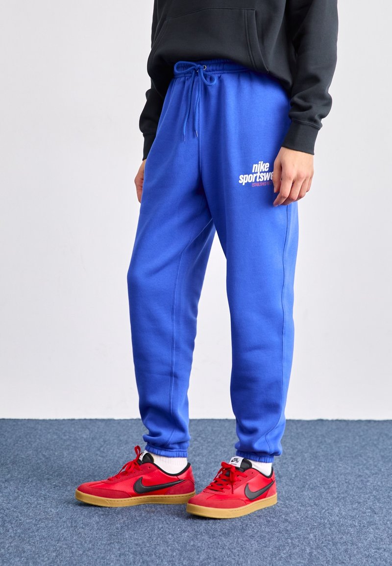 Osoba v jasně modrých sportovních kalhotách Nike a červených teniskách Nike s černým logem, stojící na modré kobercové podlaze.