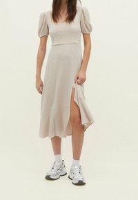 Hellbeiges Kleid mit puffigen kurzen Ärmeln, strukturiertem Stoff und einem seitlichen Schlitz, kombiniert mit silbernen Sneakers und weißen Knöchelsocken.