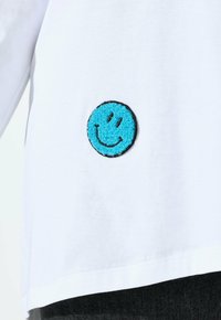 Witte shirt met een klein geborduurd blauw smiley-gezichtje bij de onderkant van de torso, gecombineerd met donkere broek.