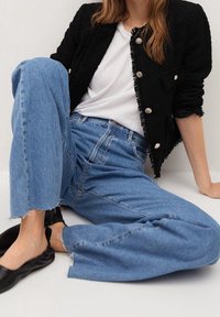 Nainen istuu lattialla mustassa tekstuurilla varustetussa takissa, jossa on hopeiset napit, valkoisessa T-paidassa, leveälahkeisissa sinisissä farkuissa ja mustissa ballerinatossuissa.