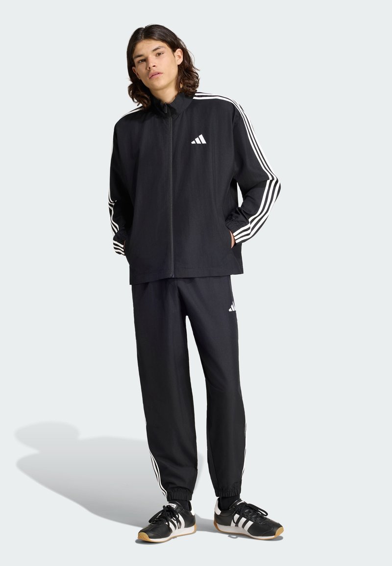 Νέος φοράει μαύρη φόρμα Adidas με λευκές ρίγες και μαύρα αθλητικά παπούτσια Adidas, στέκεται με τα χέρια στις τσέπες.