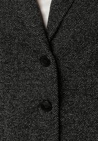 Tissu de blazer noir et blanc à pois avec une apparence texturée, comportant deux boutons noirs et un design de col classique.
