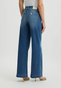 Jean en denim bleu à jambe large, avec poches arrière, décoloration légère et taille haute. Quincaillerie métallique à l'avant et détail de ourlet roulé.