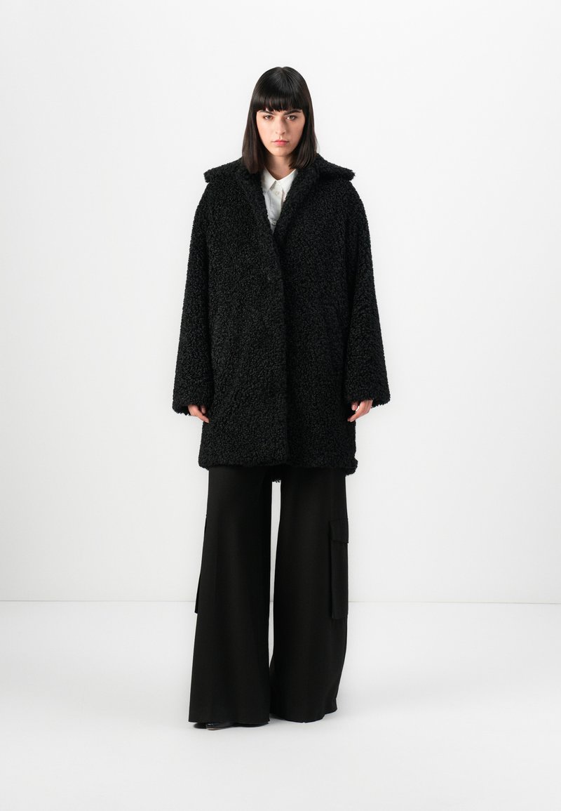 Abrigo negro de pelaje esponjoso con un acabado texturizado. Cuenta con un cuello con muescas y un corte holgado, combinado con pantalones anchos negros.
