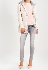 Femme portant une veste matelassée beige clair, un pull rose pâle, un jean skinny gris délavé et des chaussures à talons rose pâle debout.