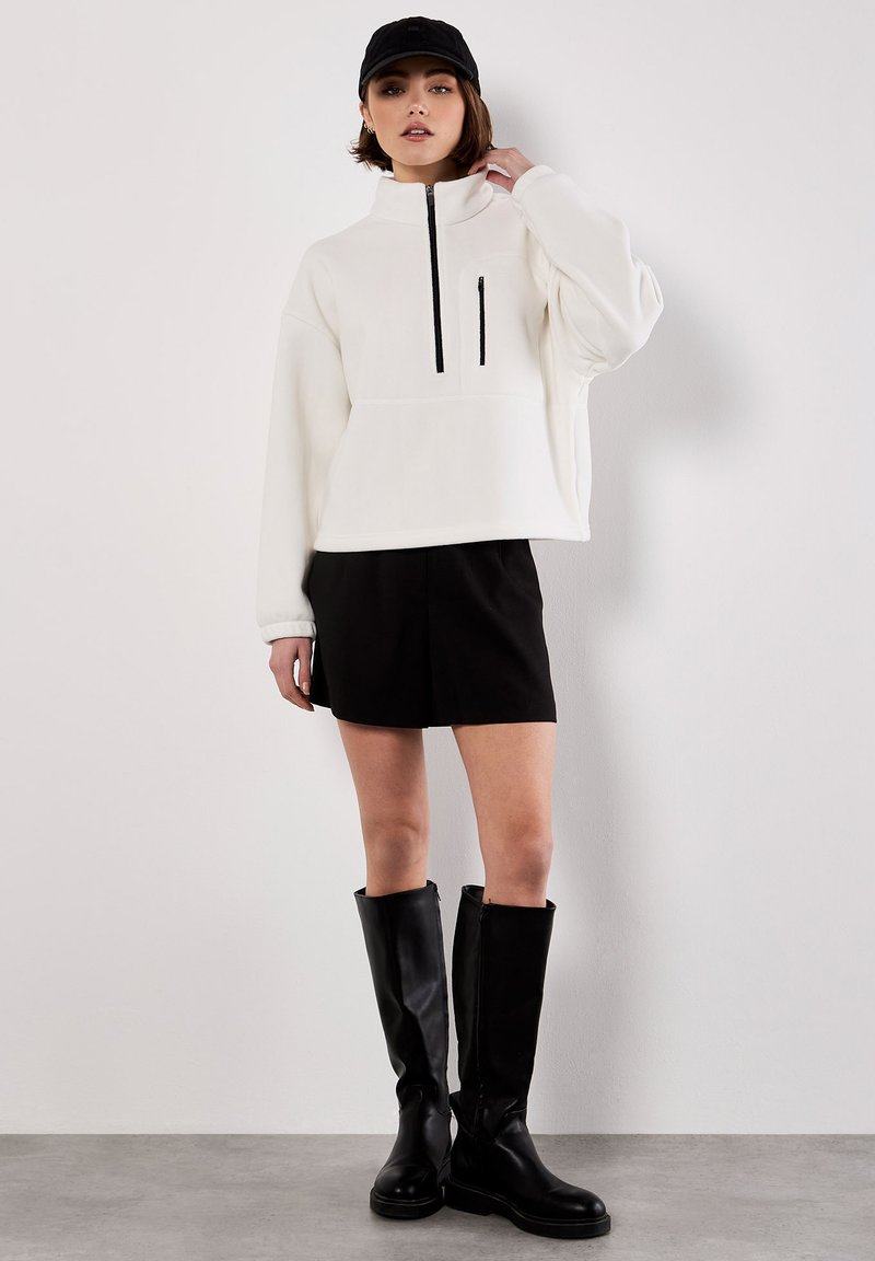 Hvid fleece-pullover med lynlås og høj krave, sort lynlåslomme, parret med sorte shorts og høje sorte læderstøvler.
