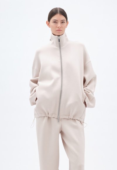 Overmaat, crèmekleurige hoodie van zacht materiaal met een hoge kraag, volledige rits en elastische zoom. Model draagt bijpassende crèmekleurige broek en staat tegen een witte achtergrond.