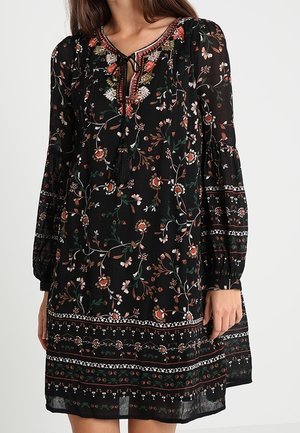 Femme portant une robe noire à fleurs avec de longues manches bouffantes et un décolleté brodé noué avec des pompons noirs.