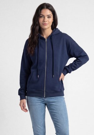 Ung kvinde med langt mørkt hår iført en marineblå hættejakke med lynlås og lyseblå jeans, står med den ene hånd på hoften.