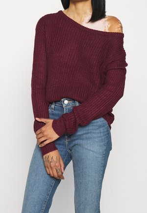 Femme avec vitiligo à l'épaule portant un pull bordeaux à épaules dénudées et un jean bleu, montrant un tatouage sur la main gauche.