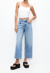 Helle blaue weit geschnittene Jeans mit fünf Taschen, hoher Taille und rohem Saum, kombiniert mit einem weißen kurz geschnittenen Tanktop und schwarzen Plateausandalen.