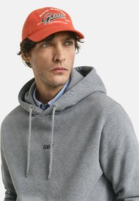 Hoodie gris avec un cordon de serrage, arborant un petit logo noir. Casquette de baseball rouge vif avec un texte blanc et vert dans un design décontracté.