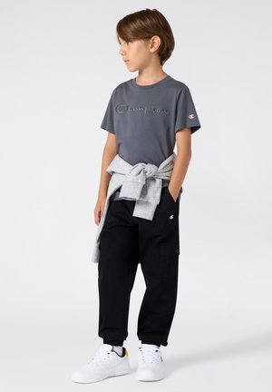 T-shirt in cotone grigio con logo "Champion" ricamato, abbinato a pantaloni cargo neri e una felpa grigia legata in vita. Sneakers bianche.