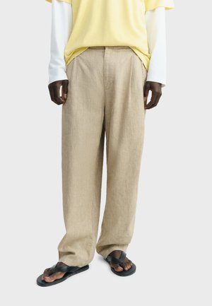 Osoba u širokim hlačama od bež lanenog platna, crnim sandalama, žutoj majici kratkih rukava preko bijele majice dugih rukava, stojeći na bijeloj pozadini.