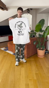 Valge suur suurusega T-särk, millel on joonistatud taimede kujutis ja tekst, koos tumedate mustriliste ööriiete pükste ja valgete tossudega, toas.