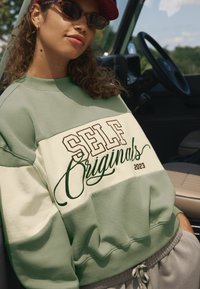 Groene en crèmekleurige oversized sweatshirt met "SELF Originals 2023" in bruine letters, voorzien van geribbelde manchetten en een relaxed fit.