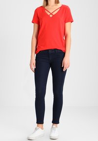 Žena v červeném krátkém rukávu topu s V-výstřihem a zkříženými ramínky, tmavě modrých skinny džínách a bílých teniskách, stojící na bílém pozadí.