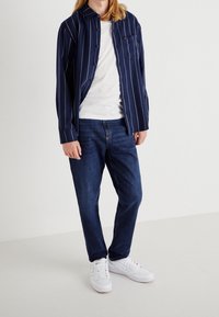 Camisa de rayas azul y blanca de manga larga, abotonada con bolsillo, usada sobre una camiseta blanca ajustada y combinada con jeans oscurecidos y zapatillas deportivas blancas.