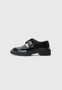 Mocasín de cuero negro brillante con una correa de hebilla plateada y suela de goma gruesa con textura, mostrado de perfil sobre un fondo blanco.