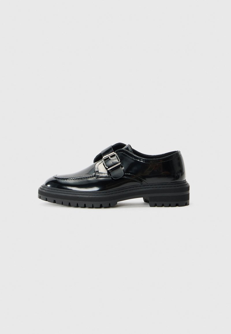 Mocasín de cuero negro brillante con una correa de hebilla plateada y suela de goma gruesa con textura, mostrado de perfil sobre un fondo blanco.