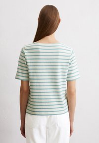 Gestreiftes kurzärmliges Oberteil in hellem Beige mit türkisfarbenen horizontalen Streifen. Aus weichem Baumwollmaterial, Rundhalsausschnitt und lockerer Passform. Weiße Hose sichtbar.