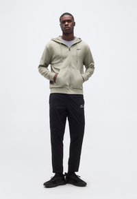 Νεαρός άνδρας στέκεται με τα χέρια στις τσέπες, φορώντας ανοιχτό πράσινο φούτερ με φερμουάρ, μαύρο παντελόνι Nike και μαύρα αθλητικά παπούτσια σε λευκό φόντο.