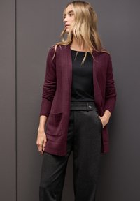 Cárdigan burdeos con apertura frontal, puños acanalados y dos bolsillos delanteros, combinado con una blusa negra y pantalones grises a medida. Textura suave.