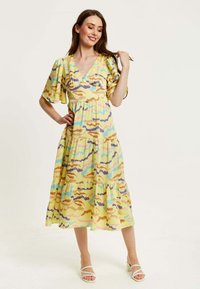 Yellow patterned dress featuring a V-neck, short puff sleeves, tiered design, and multicolored abstract swirls; worn with white sandals.
