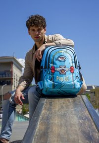 Mochila azul con un gráfico de ojo, herrajes rojos y cierre de cremallera. Equipado con bolsillos laterales de malla y un diseño estructurado.