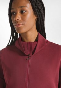 Giacca zip-up bordeaux con colletto alto, tessuto liscio e zip metallica. Il design presenta una silhouette elegante e aderente.