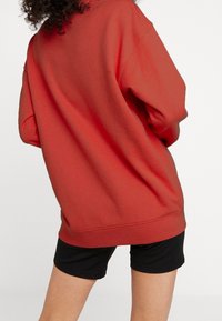 Röd oversized sweatshirt med en slät textur och rundad halsringning, kombinerad med svarta shorts. Visas bakifrån och framhäver tröjans form.