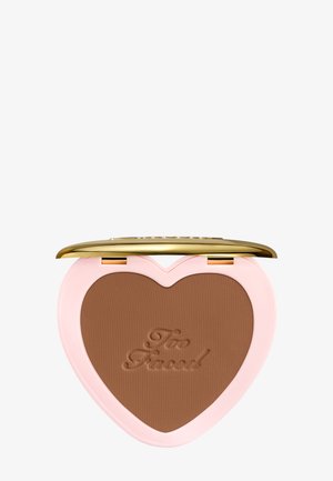 Herzförmiges Kompaktpuder mit braunem Puder-Makeup, geprägt mit "Too Faced", pinkfarbene Hülle und teilweise geöffneter goldener Deckel vor weißem Hintergrund.
