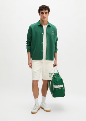 Jeune homme portant une veste verte à boutons, un short blanc avec le numéro "67" imprimé, des baskets blanches, tenant un sac de sport vert avec le logo "Marc O’Polo".
