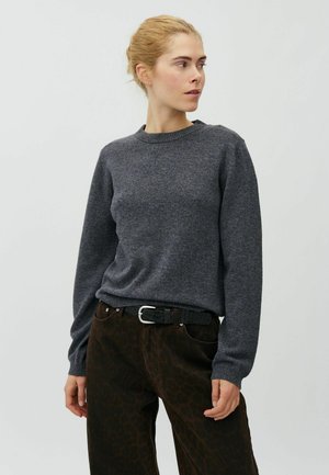 Femme aux cheveux blonds portant un pull gris foncé à manches longues et un pantalon foncé à motifs, regardant sur le côté sur un fond uni.