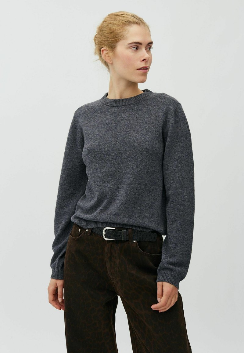 Femme aux cheveux blonds portant un pull gris foncé à manches longues et un pantalon foncé à motifs, regardant sur le côté sur un fond uni.