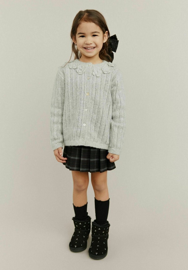 Vaaleanharmaa kaarrokepaita, jossa on kukkakoristeita, mustat ruudulliset hame, polven yli ulottuvat sukat ja mustat saappaat valkoisilla yksityiskohdilla.