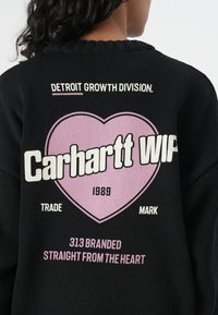 Zwarte gebreide trui met een groot roze hartontwerp. De tekst bevat "Carhartt WIP," "Detroit Growth Division," en "1989." De textuur is geribbeld.