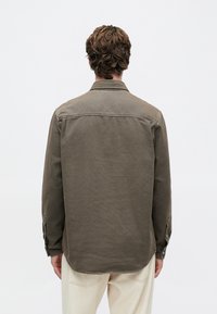 Hombre con cabello castaño ondulado, vestido con una camiseta de manga larga verde oliva y pantalones beige claros, de pie frente a un fondo blanco liso.