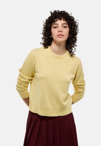 Pull en tricot jaune à manches longues, col et ourlet côtelés, associé à une jupe plissée bordeaux pour un contraste de couleur et de texture.