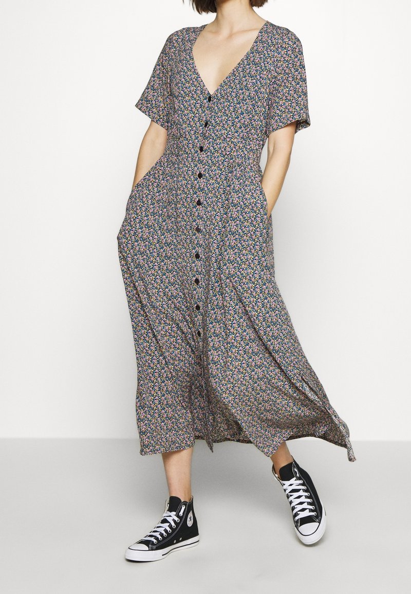 Robe maxi fleurie à manches courtes, encolure en V, taille cintrée et poches latérales. Comprend des boutons noirs et un tissu léger à motifs.