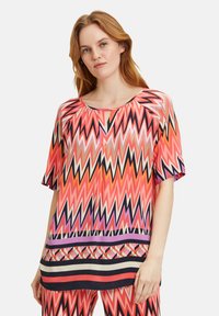 Blouse multicolore avec un motif en zigzag rose, orange, noir et blanc. Manches courtes et col rond avec un détail en trou de serrure.