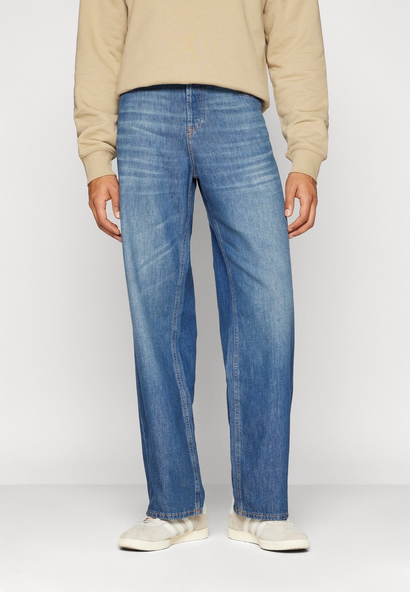 Personne portant un jean bleu droit, un sweat-shirt beige et des baskets blanches, debout devant un fond uni.