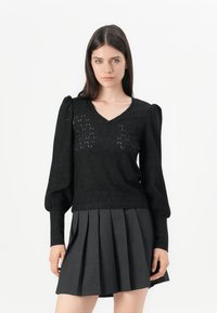 Maglione lavorato a maglia nero con scollo a V, maniche a palloncino e motivo a diamante aperto. Abbinato a una gonna plissettata grigia.
