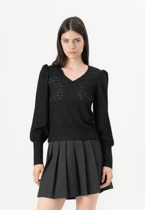 Maglione lavorato a maglia nero con scollo a V, maniche a palloncino e motivo a diamante aperto. Abbinato a una gonna plissettata grigia.