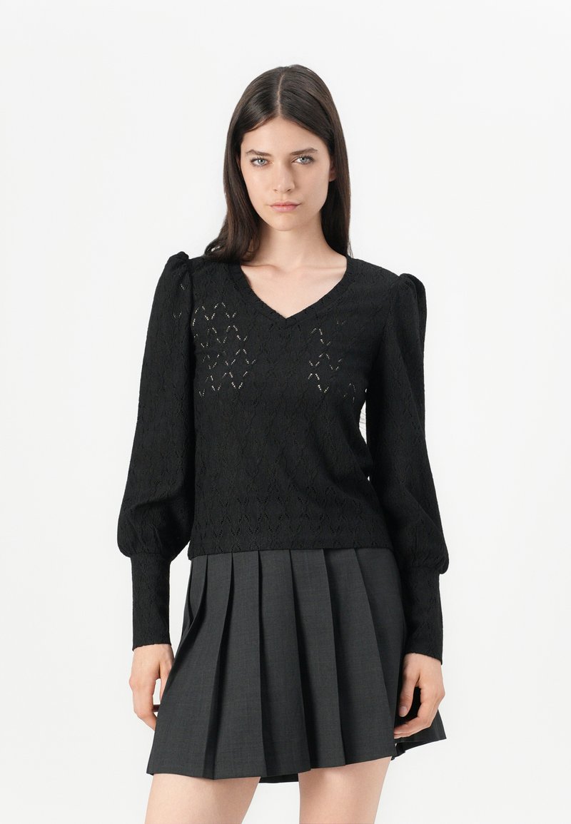 Maglione lavorato a maglia nero con scollo a V, maniche a palloncino e motivo a diamante aperto. Abbinato a una gonna plissettata grigia.