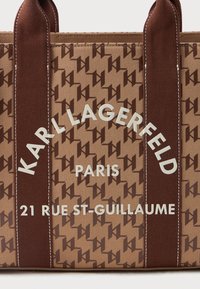 Bolso de mano marrón con un patrón de logotipo marrón oscuro repetido, asas marrones con costuras blancas y texto blanco "Karl Lagerfeld Paris 21 Rue St-Guillaume."