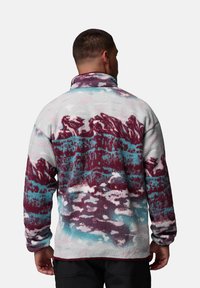 Fleece-Pullover mit hohem Kragen, der ein mehrfarbiges Bergpanorama-Muster in Burgunderrot, Petrol und Weiß vor einem hellen Hintergrund zeigt.